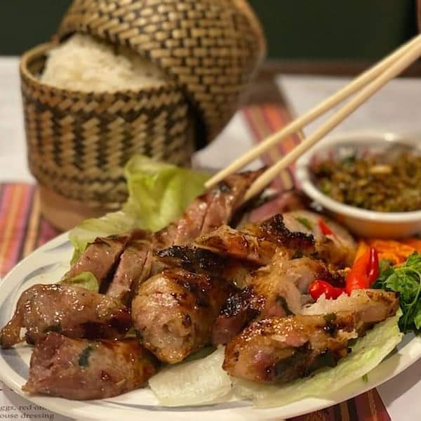 Grilled skewered meat on a plate with sticky rice in a woven basket and a side of sauce.