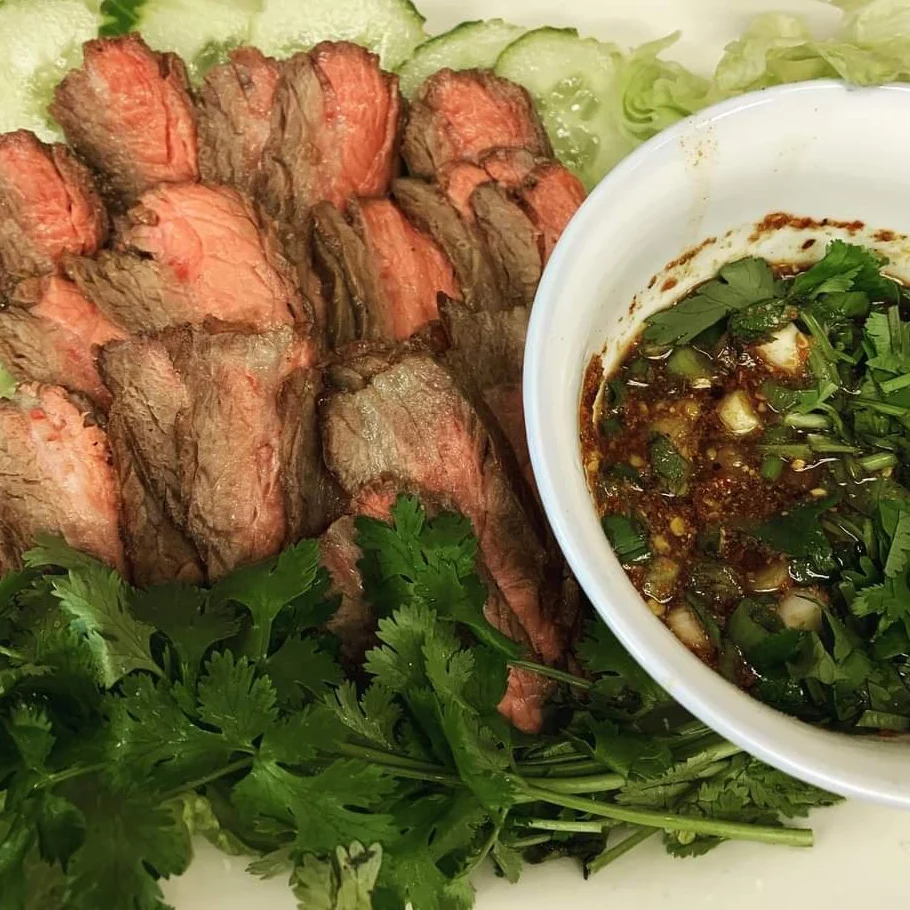 Grilled skewered meat on a plate with sticky rice in a woven basket and a side of sauce.