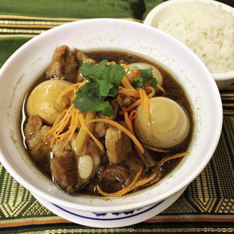 Bowl of stew with meat, eggs, and carrots, garnished with cilantro, next to rice on a green mat.